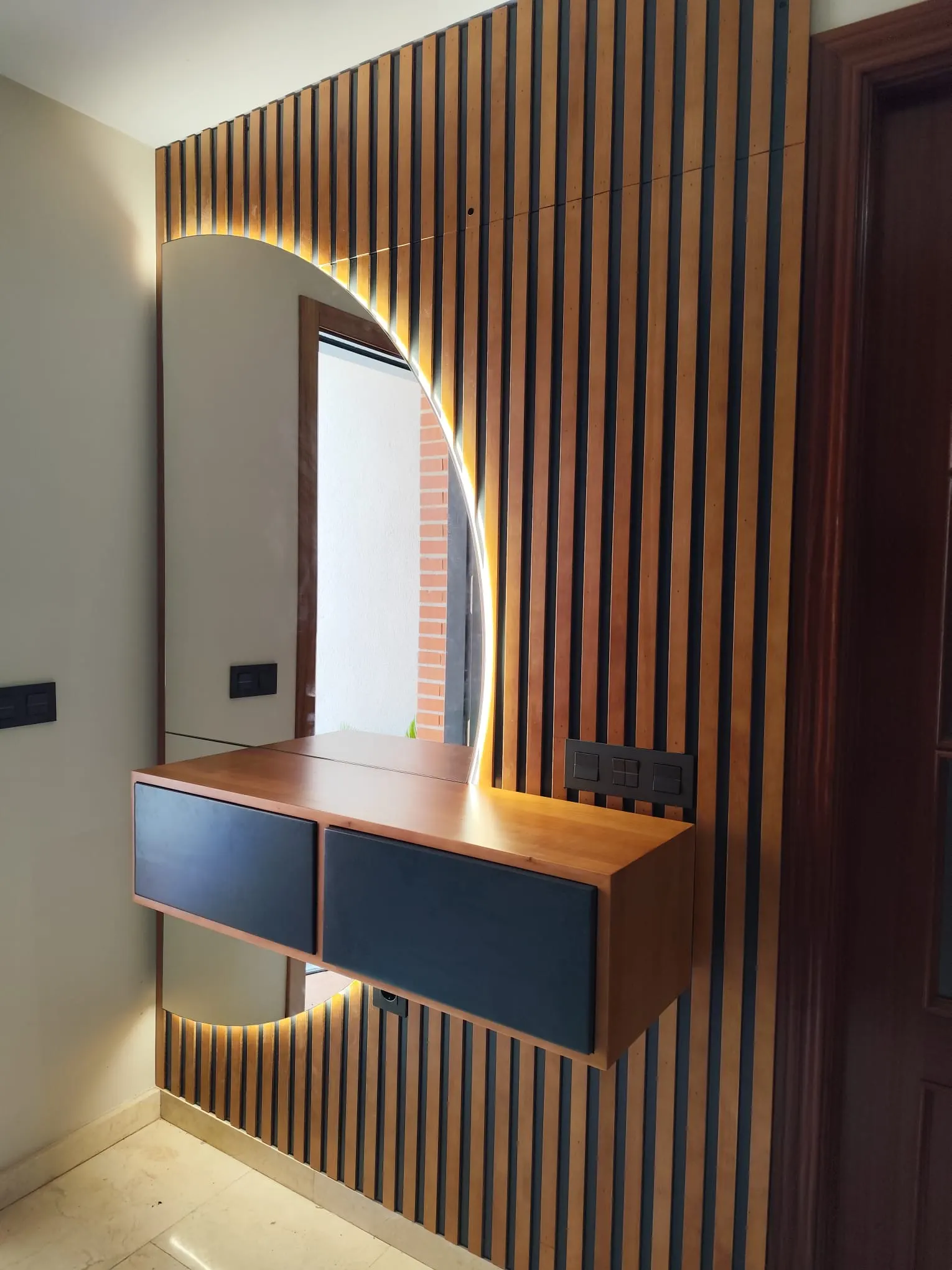 Recibidor con pared de listones de madera, espejo semicircular retroiluminado y mueble colgado de dos cajones